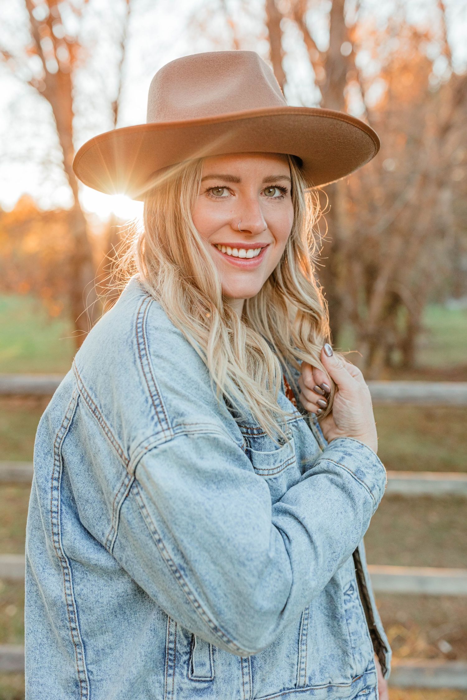 Woman in denim jacket and brown hat poses outdoors during golden hour with autumn trees in background.