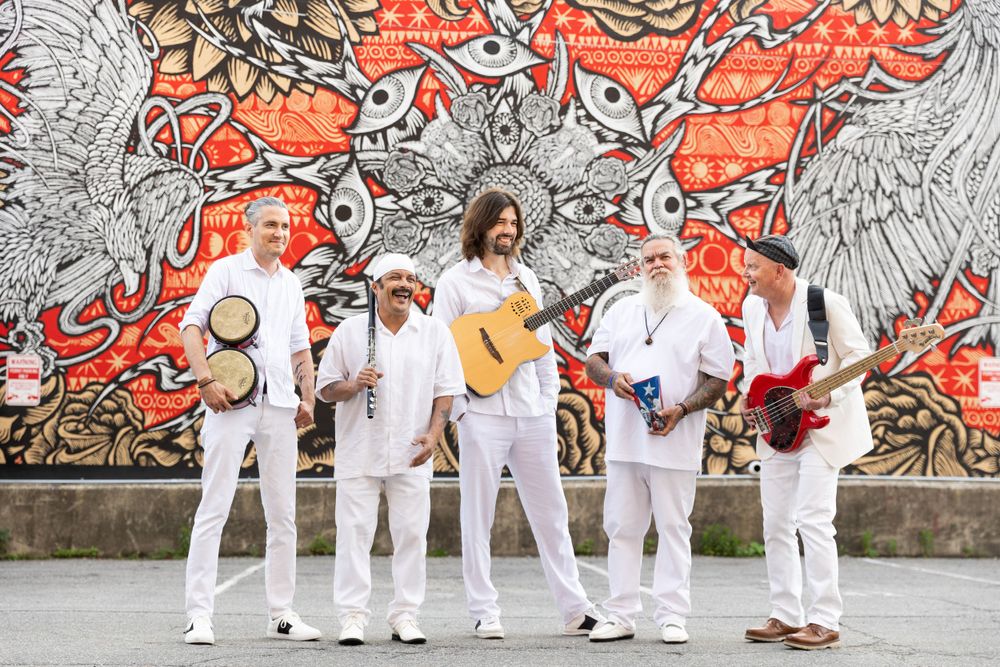 Musicians in white outfits performing against colorful graffiti wall backdrop.