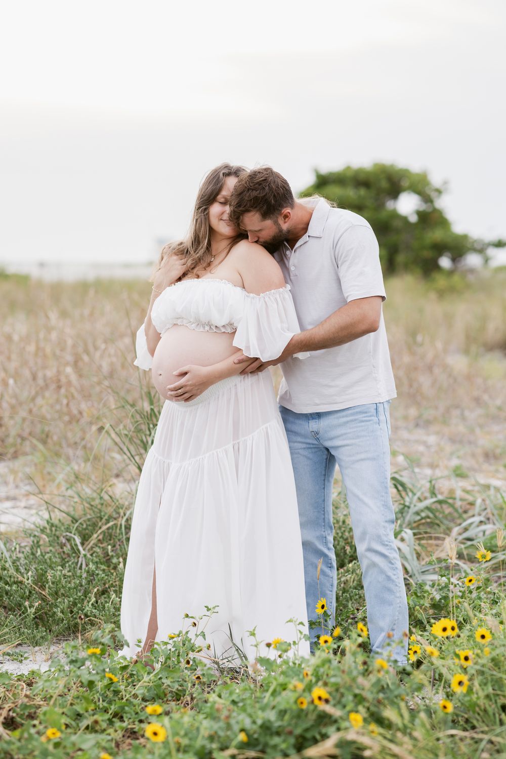 Man in jeans kisses expectant woman wearing white outfit on shoulder standing in daisies and coastal grass. Salisbury maternity photographer