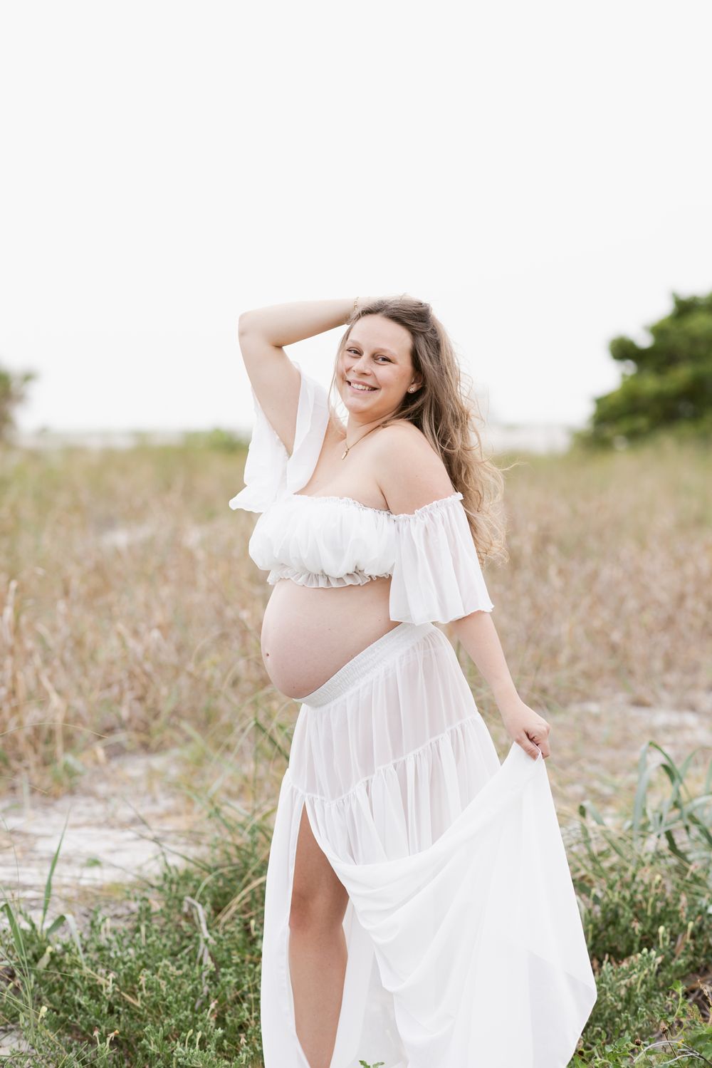 Happy expectant mother in white sheer dress poses in coastal grass for maternity photos. Southern Maine maternity photographer