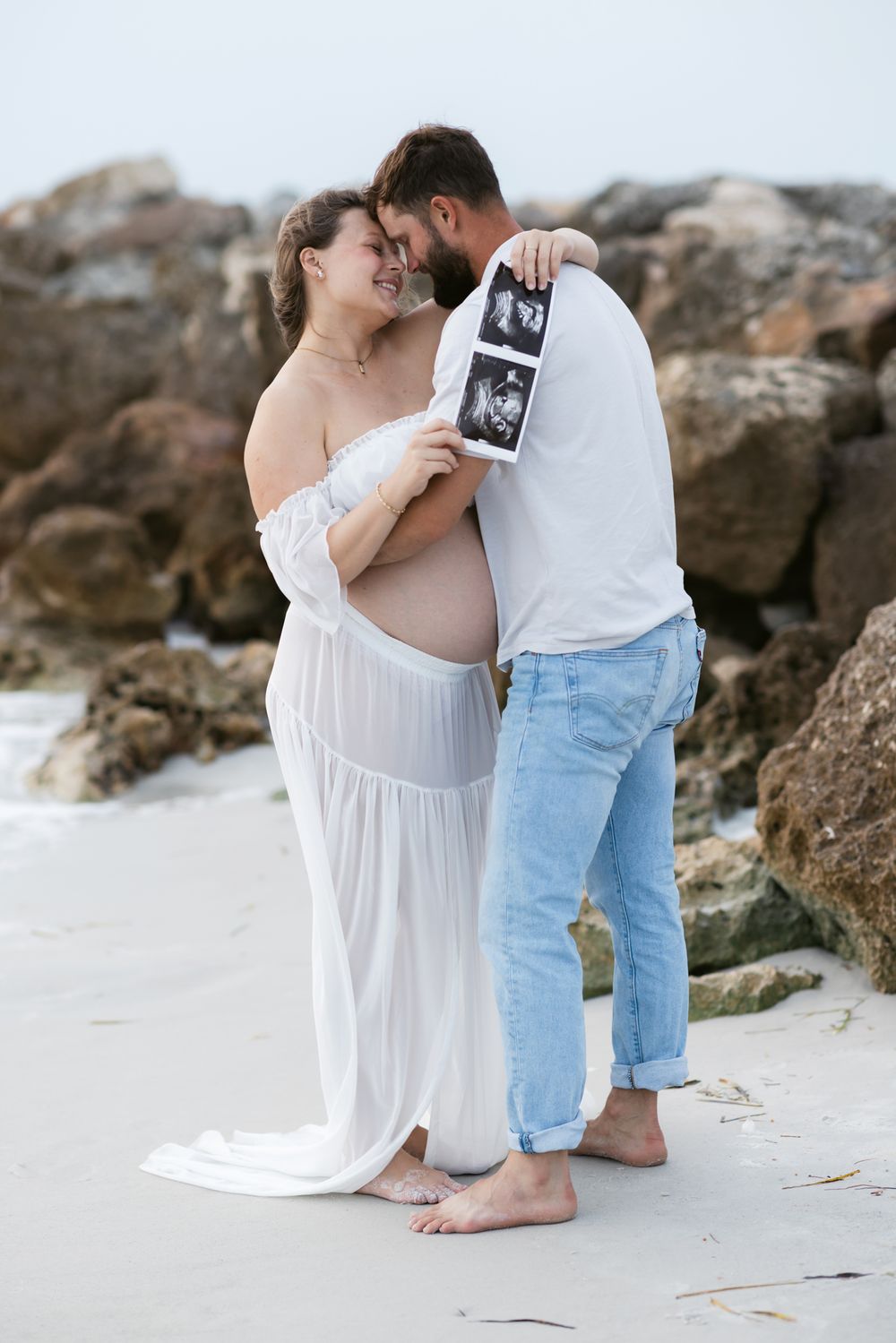 Expectant mother in sheer skirt and man in jeans embrace holding an ultrasound photo in front of brown coastal rocks for maternity photos.