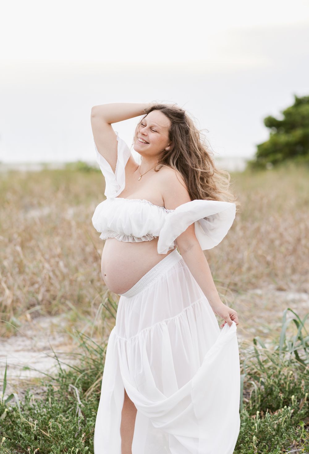 Exuberant expectant mother wears a sheer maternity dress posing in coastal grass. Rye York Newburyport maternity photographer
