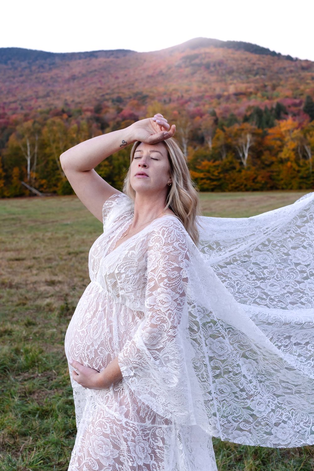 Expectant mother in white lace dress poses dramatically in open field with mountain in background. North Conway maternity photographer.