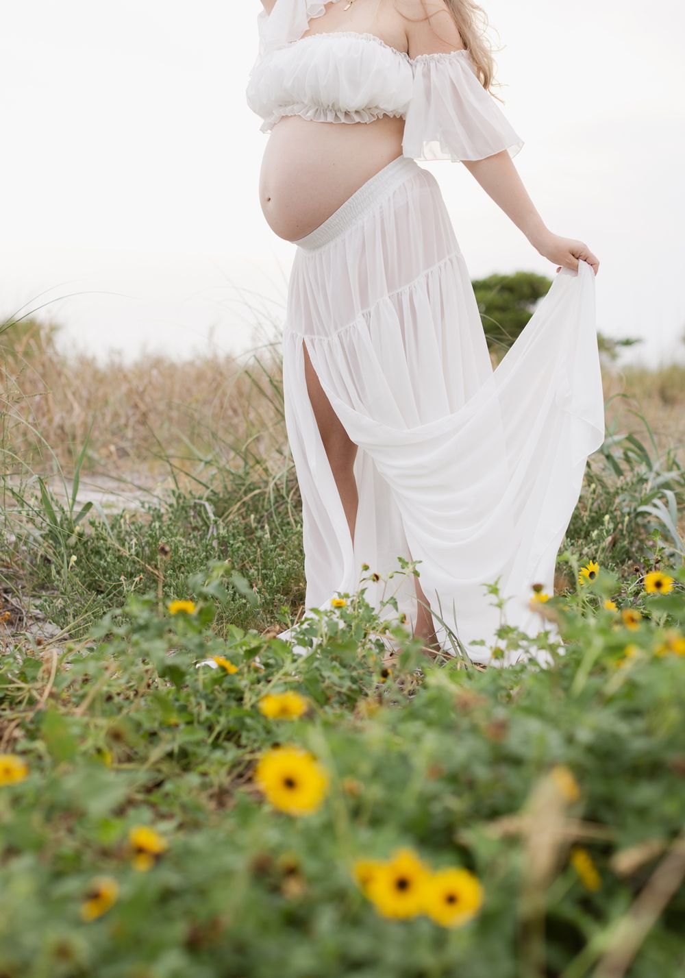 Out of focus cropped expectant mother reveals her bump posing in daisies and coastal grass. Lakes Region, Concord maternity photographer
