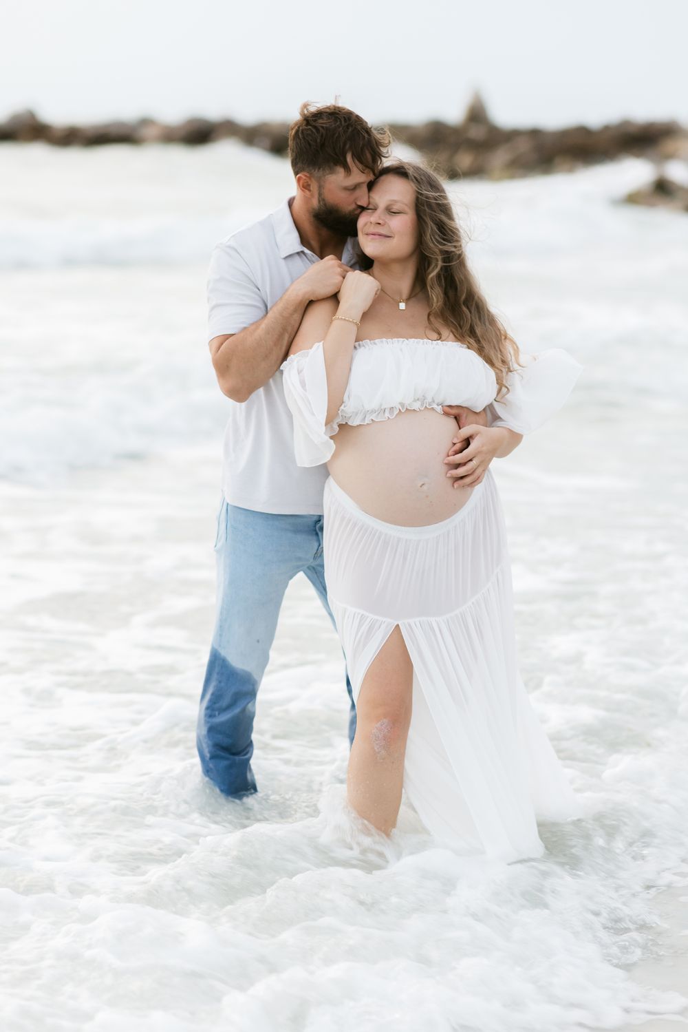 Man embraces woman from behind in ruffled cropped top and white skirt standing in shallow ocean water. North Shore maternity photographer.