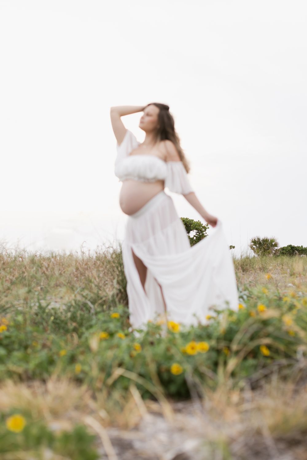 Outline of blurry expectant figure poses in daisies and coastal grass on cloudy day for maternity photos. Seacoast NH maternity photographer