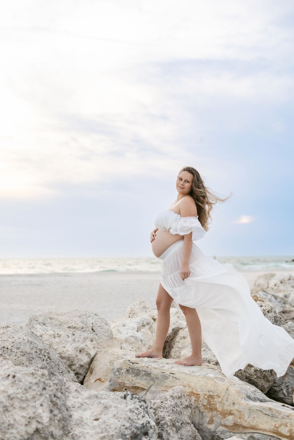 Expectant mother in sheer white dress and cropped top stands on light large coastal rocks during cloudy sunset. Southern Maine maternity.