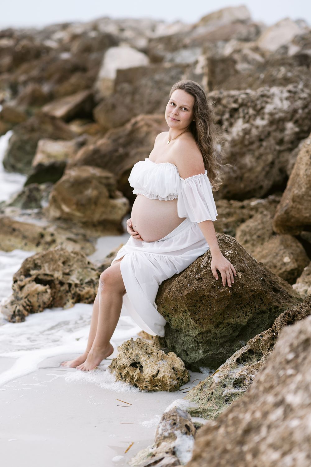 Expectant mother wears sheer white outfit sitting on brown coastal rocks for maternity photos. Concord Bedford maternity photographer