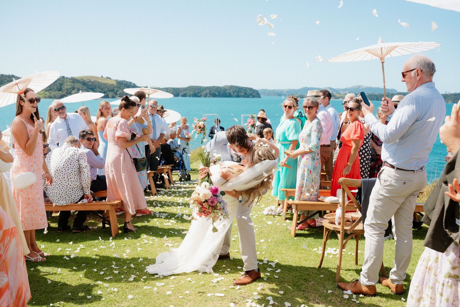 Guests celebrate during an outdoor wedding ceremony on a grassy lawn overlooking the ocean on a sunny day.