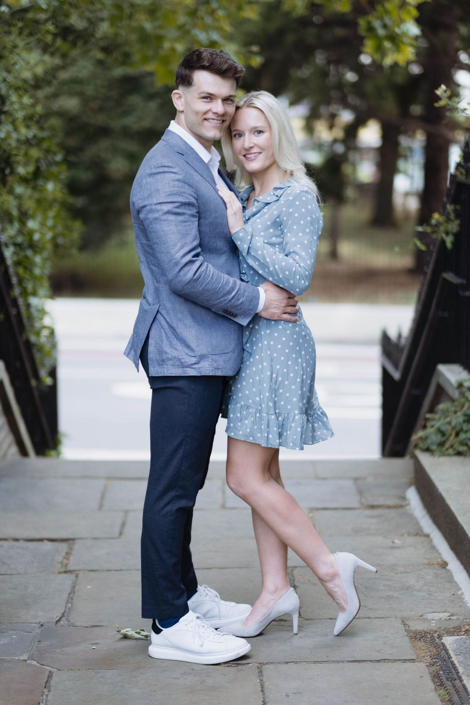 Man in a suit and sneakers holding a woman in a blue dress and high heels outdoors near a street and greenery