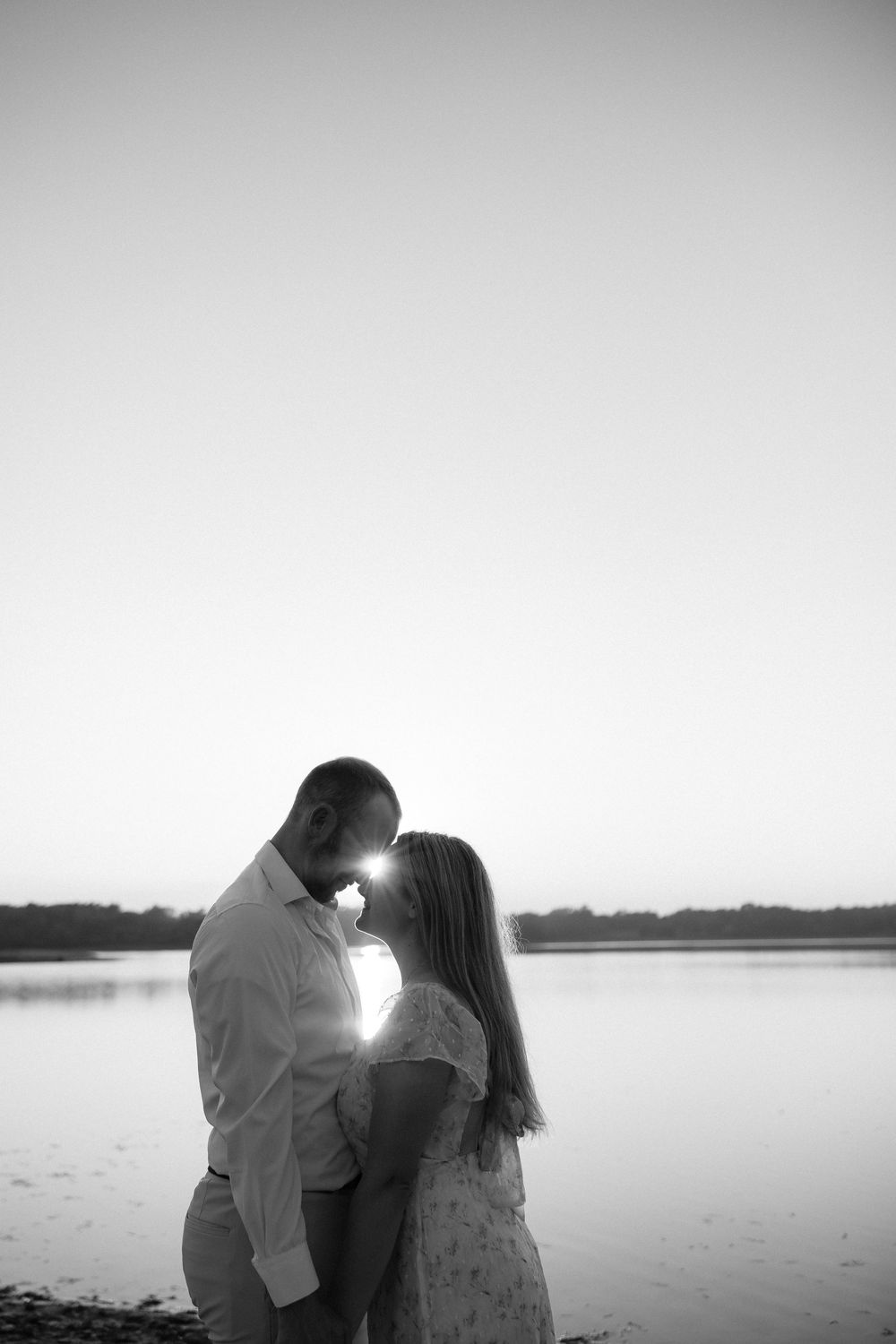 Black and white photo of a couple sharing a romantic moment by a lake at sunset, with sun flare between them.