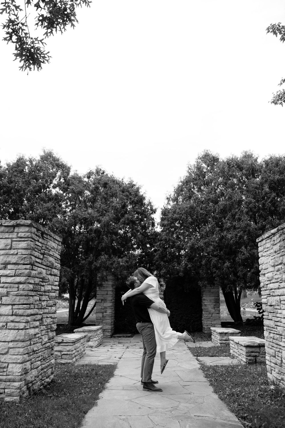 A loving embrace between two people on a stone pathway framed by stone pillars and trees in black and white.