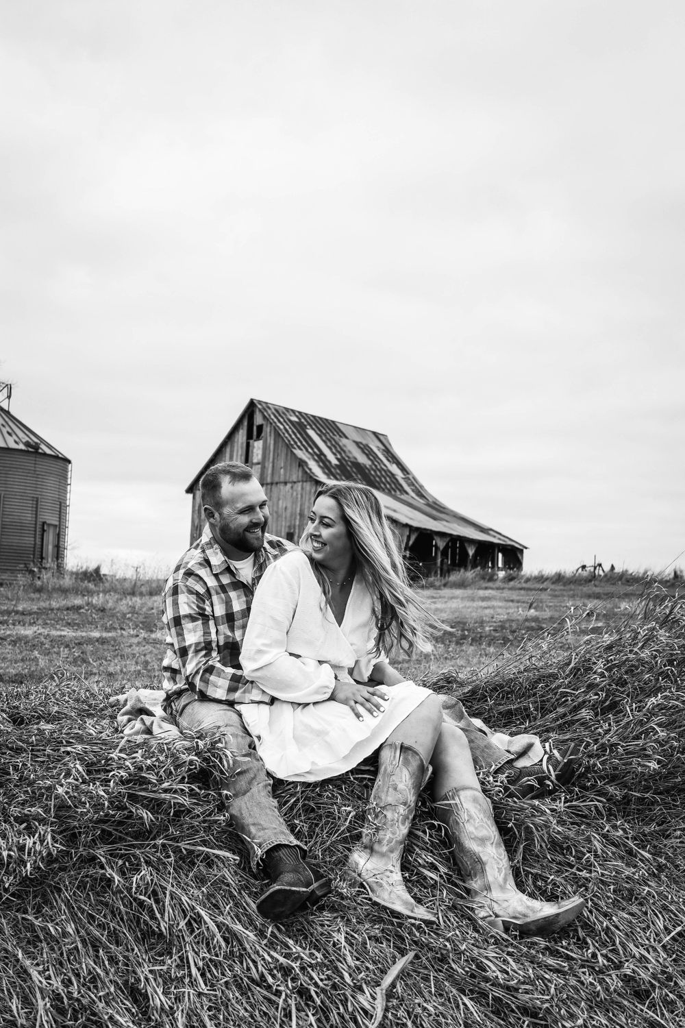 A couple sits together in rustic boots near an old weathered barn during a countryside photoshoot.