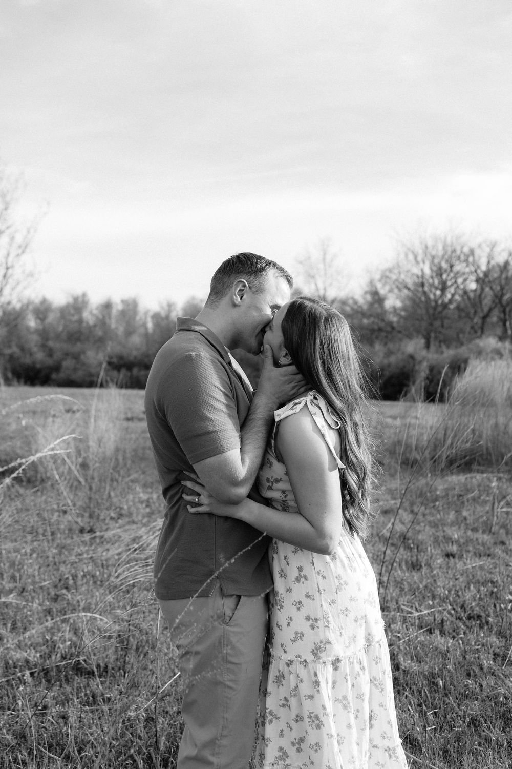 Black and white photo of a couple sharing a romantic kiss in a field with trees in the background.