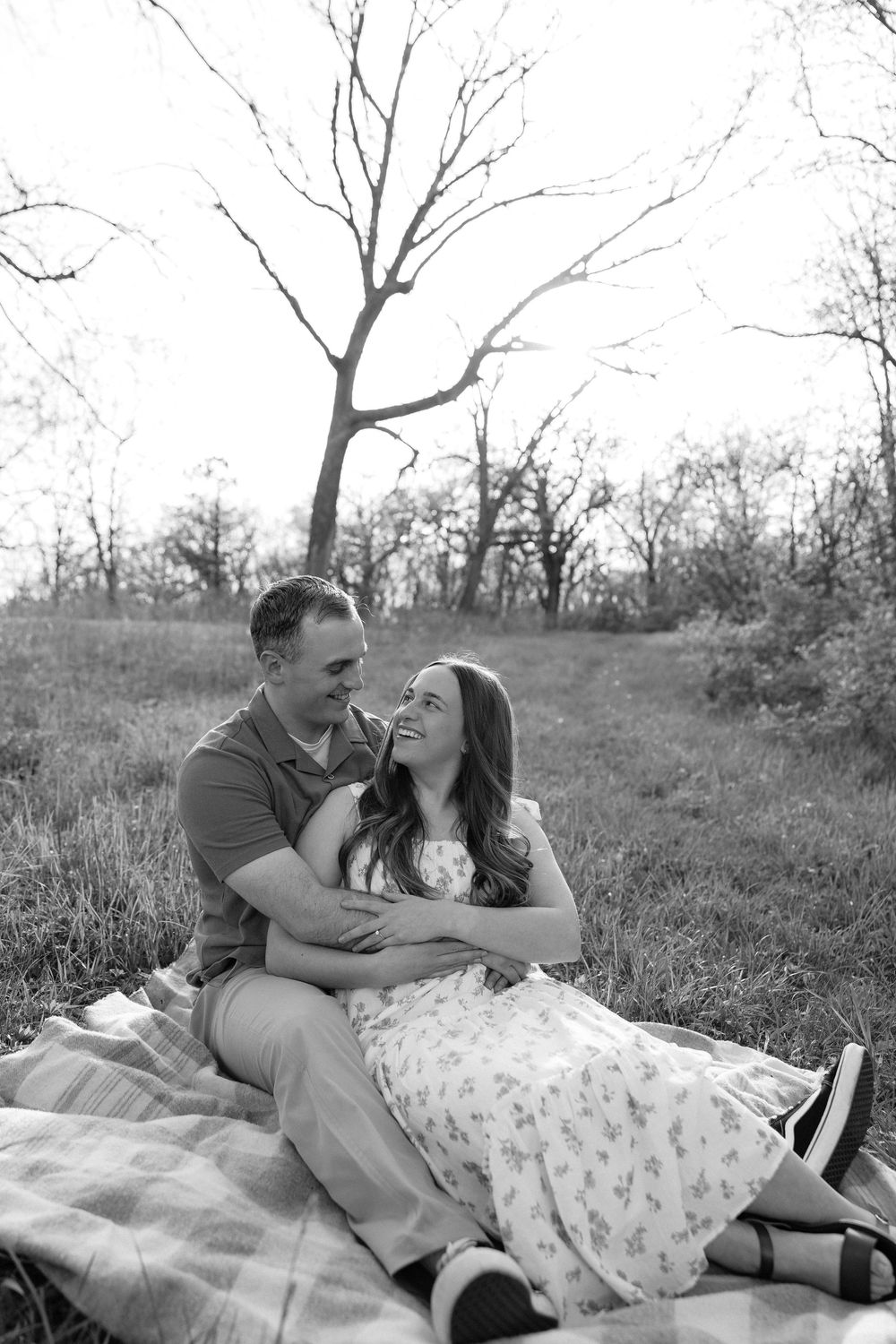 Black and white photo of a couple sitting on a blanket in a field, laughing and embracing during an outdoor engagement session.