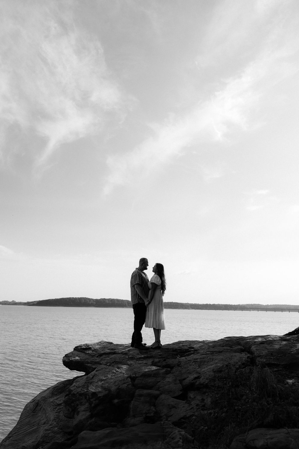 A romantic silhouette of a couple stands on a rocky cliff overlooking a serene lake under cloudy skies.