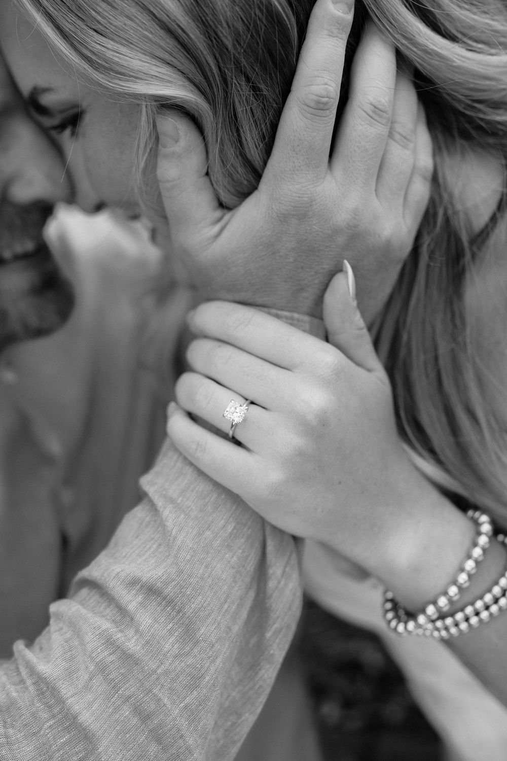 Black and white photo of couple sharing intimate moment, woman displaying diamond engagement ring and pearl bracelet.