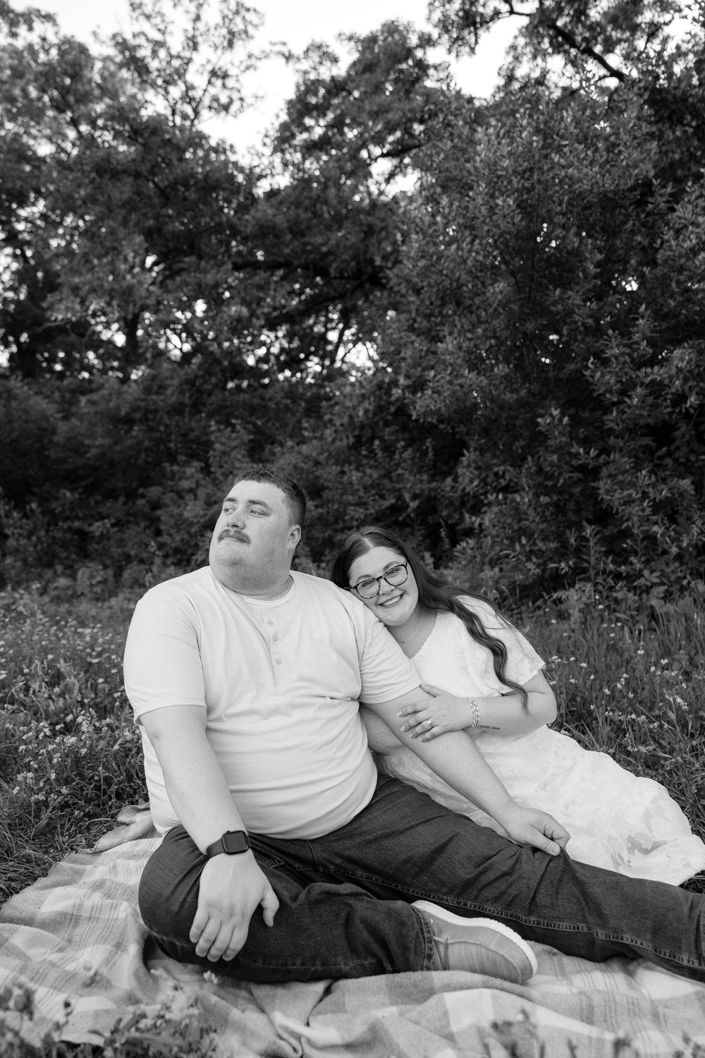 Black and white photo of a couple sitting on a blanket outdoors, smiling and relaxing together in a lush green setting.