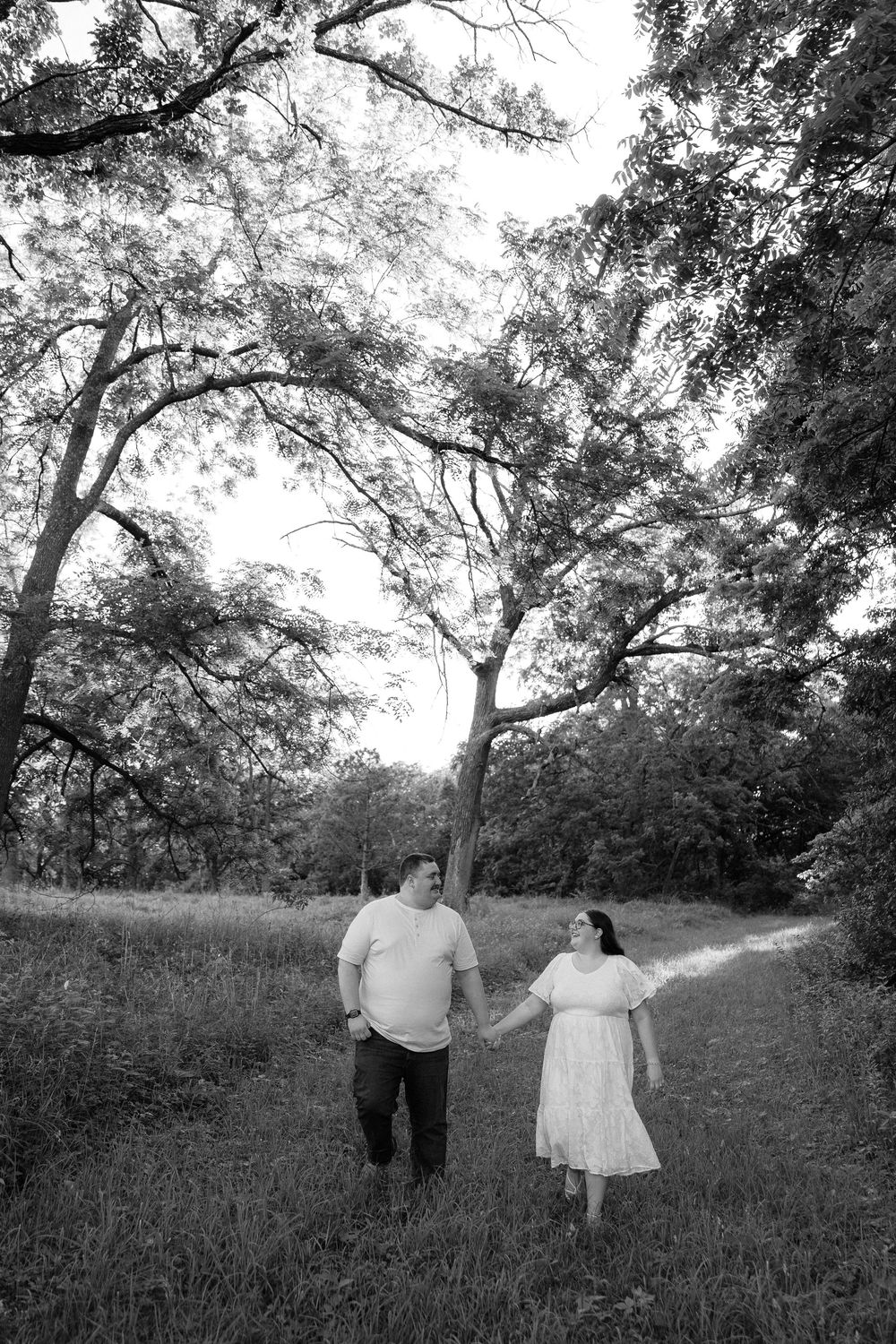 Black and white photo of a couple holding hands on a winding path surrounded by tall trees in a lush outdoor setting.
