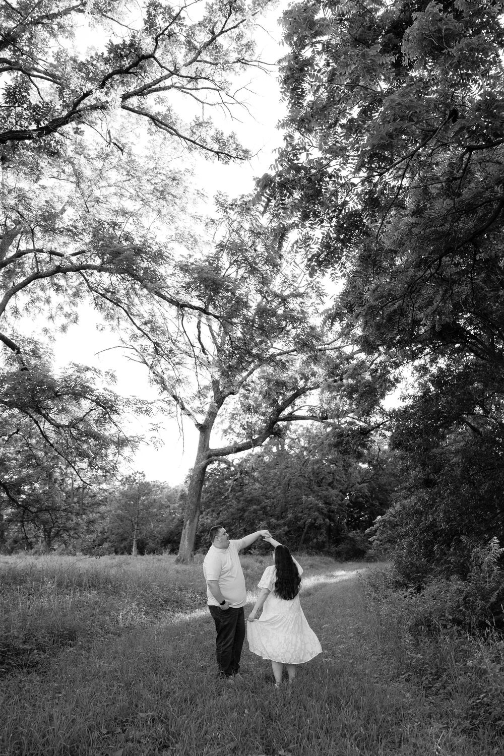 Black and white photo of a couple dancing on a tree-lined path in a lush park setting.