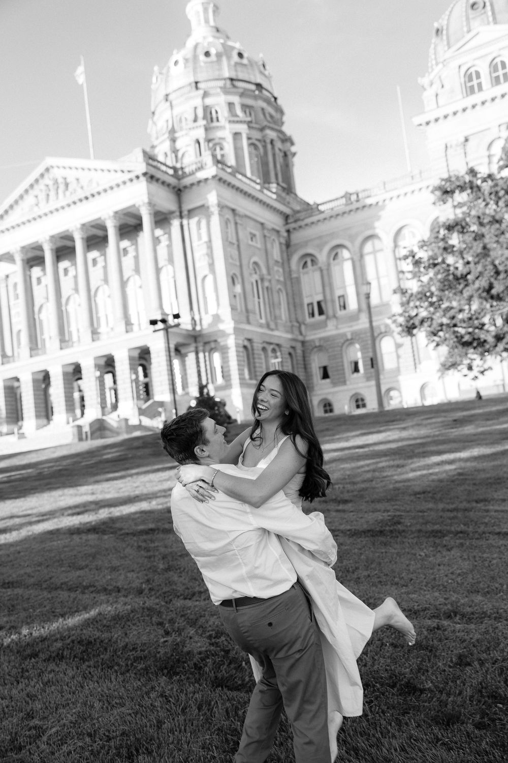 Black and white photo of a couple embracing and laughing in front of a grand capitol building on a sunny day.