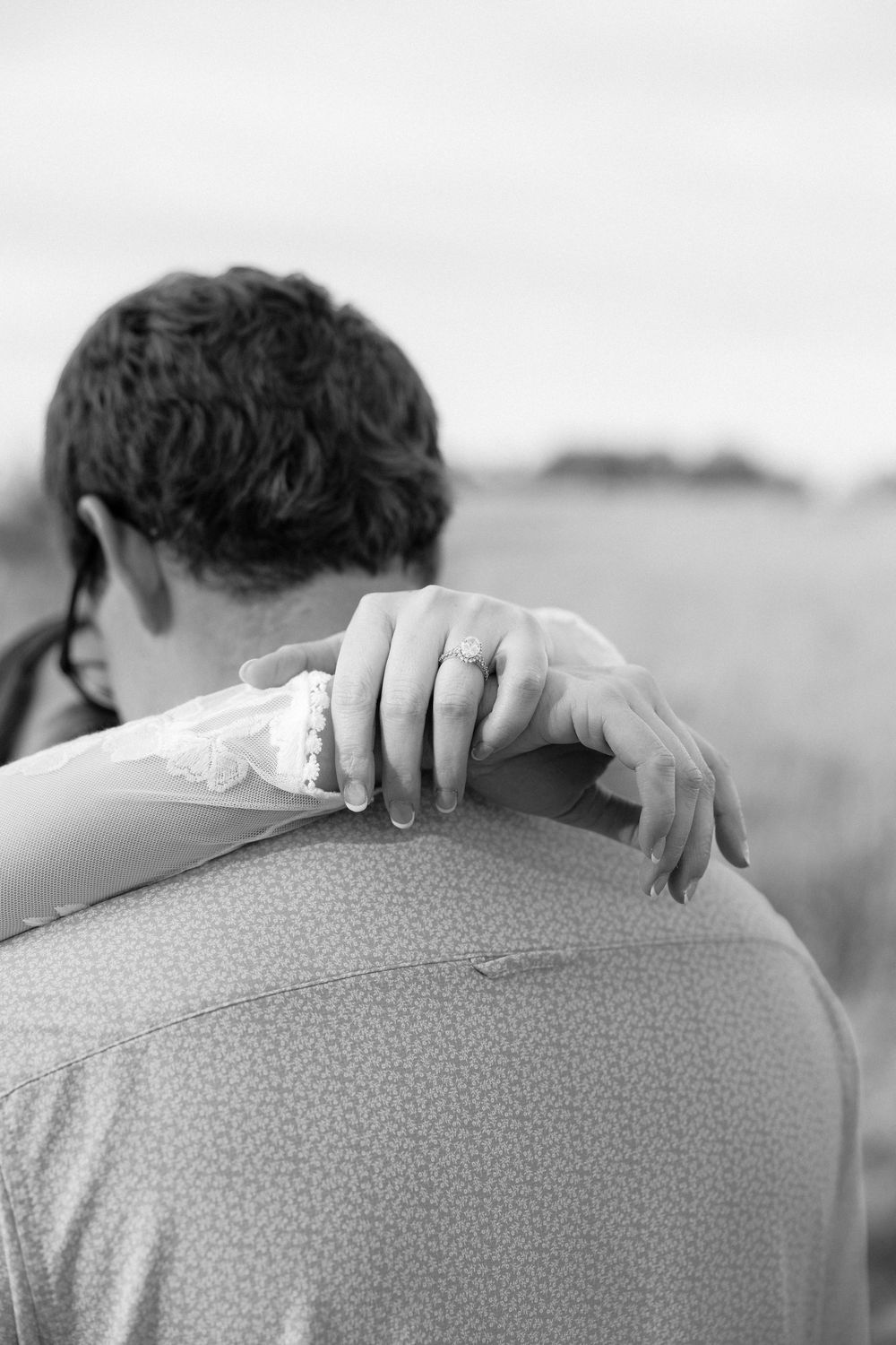 Black and white photo of a bride resting her hand on groom's shoulder, showing engagement ring, in a field.