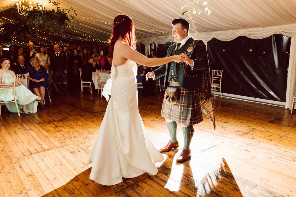 Wedding party dances on wooden floors under elegant white tent draping and chandeliers.