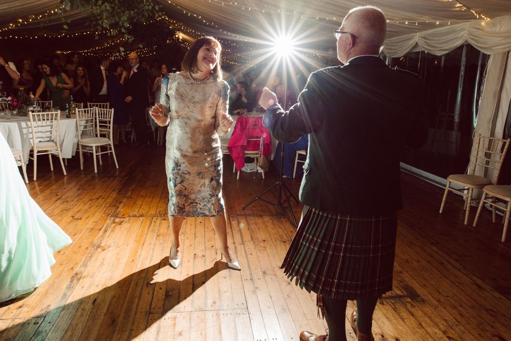 Wedding guests dance on wooden floors under elegant white tent draping and chandeliers.