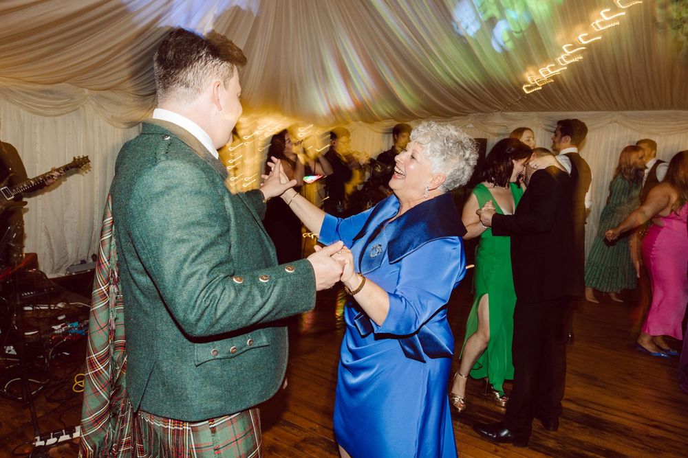 Dancing guests at a Scottish wedding with traditional kilts and formal attire under decorative lighting.
