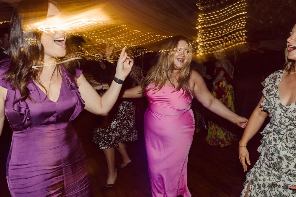 Wedding guests dancing in colorful formal dresses under warm tent lighting.