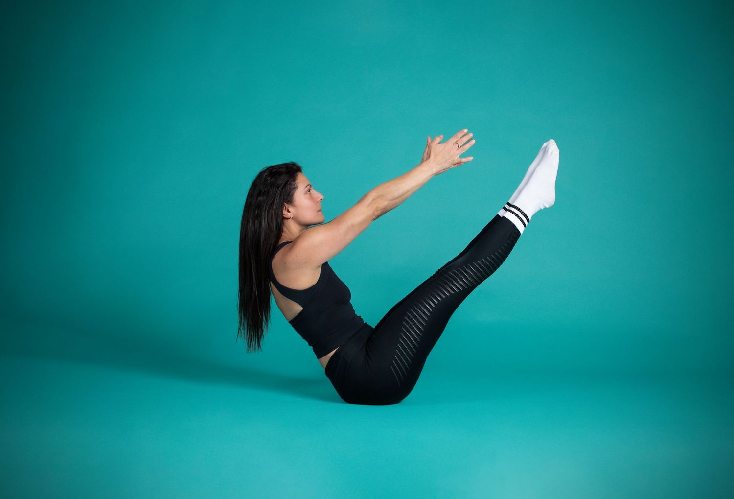 Against teal backdrop, person in black workout attire performs V-sit core exercise on studio floor.