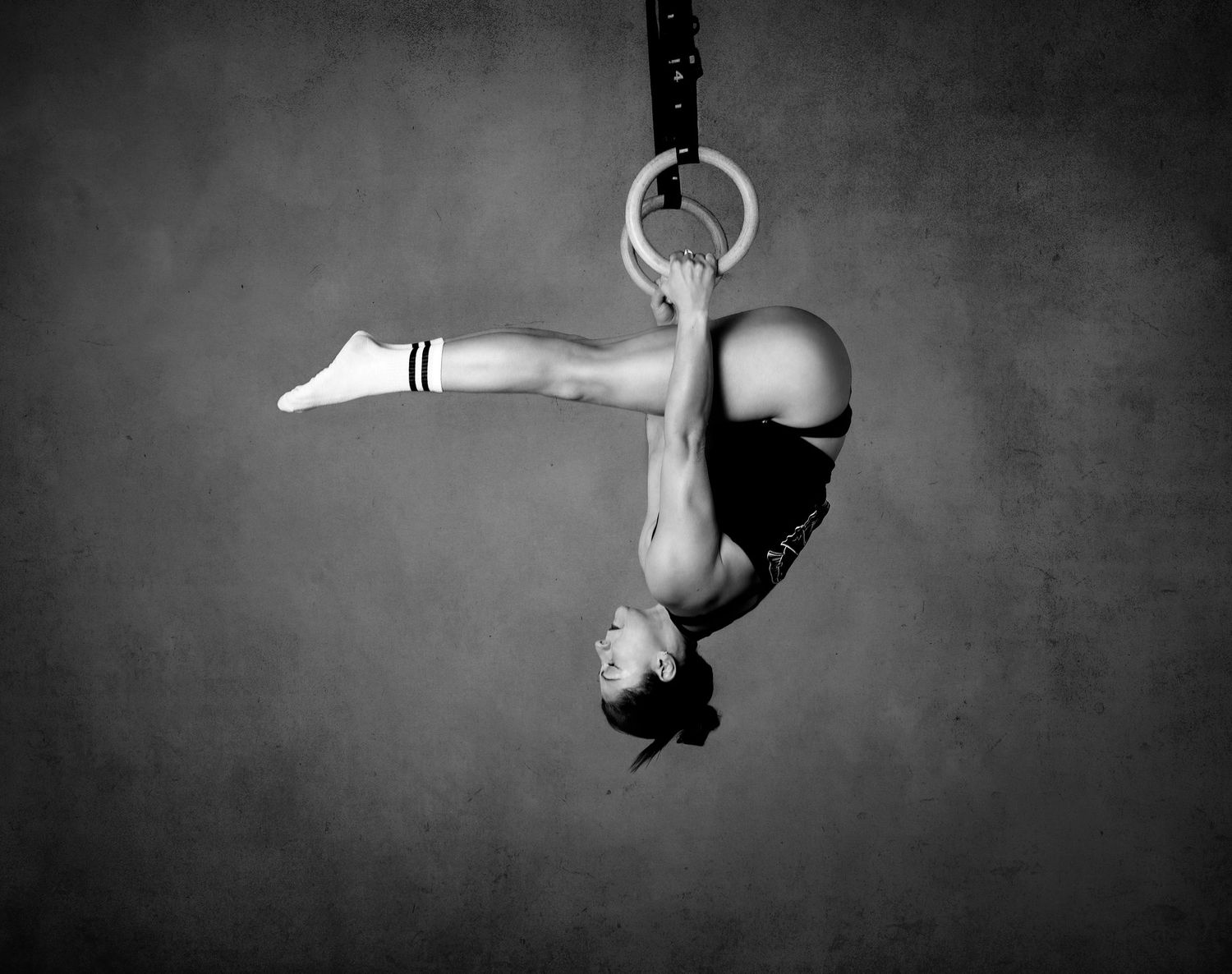 A gymnast hangs upside down from rings in a dramatic black and white artistic photograph.