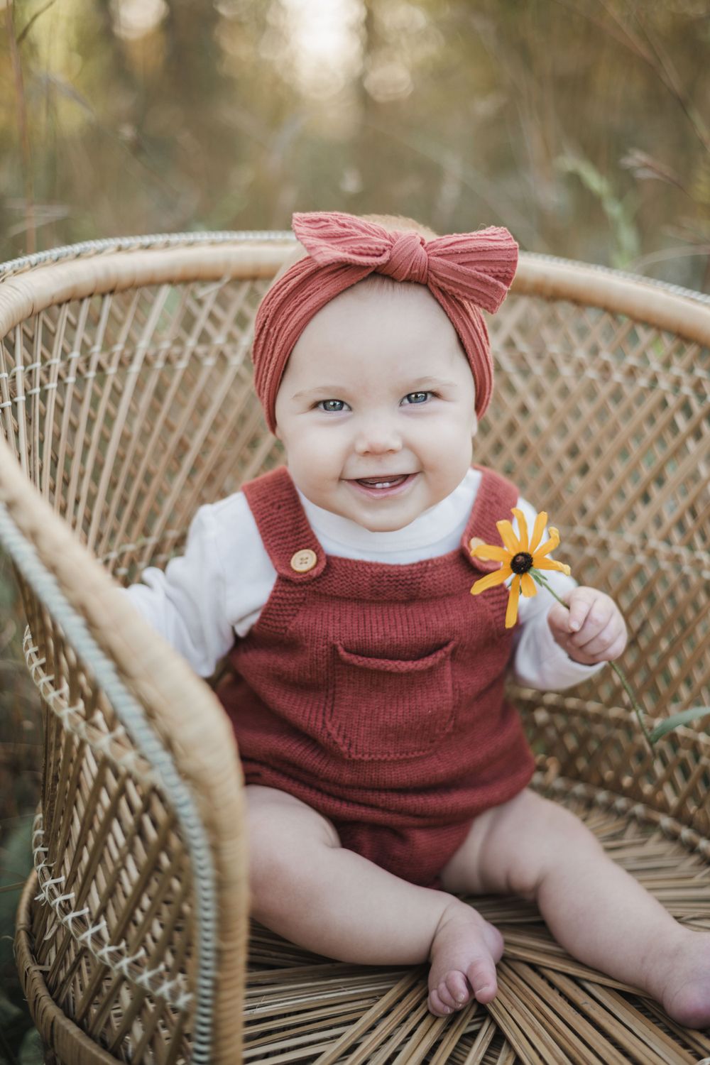 Baby in burgundy romper with bow headband sitting in wicker chair outdoors.