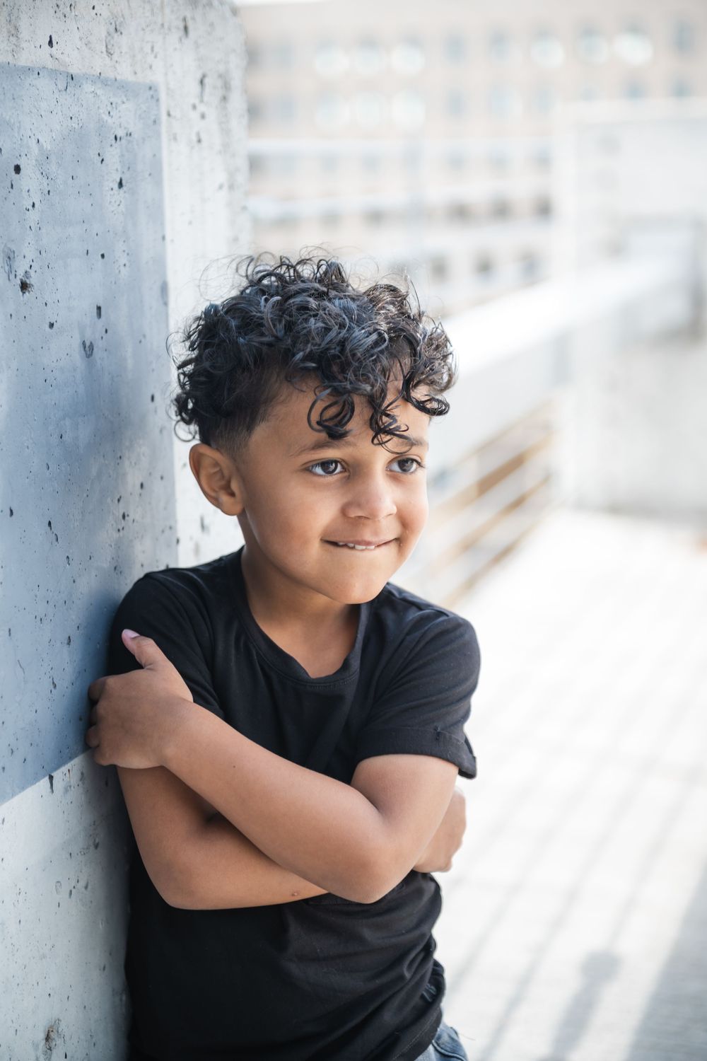 Young boy with curly hair in black t-shirt smiling with arms crossed, leaning against concrete wall outdoors.