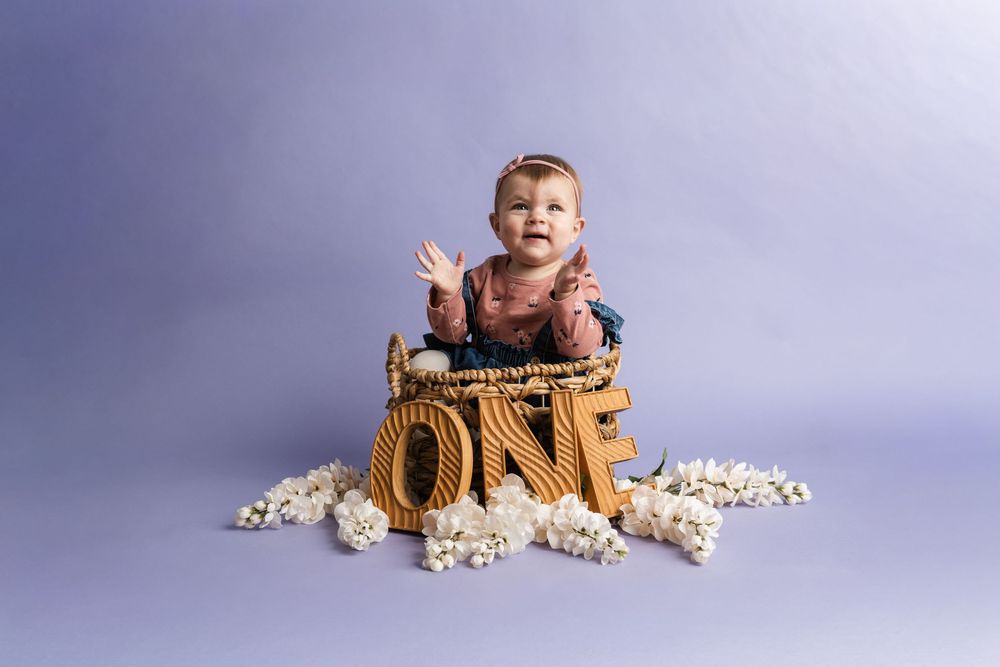 A small child sits in a vintage baseball glove surrounded by scattered popcorn against a purple backdrop.