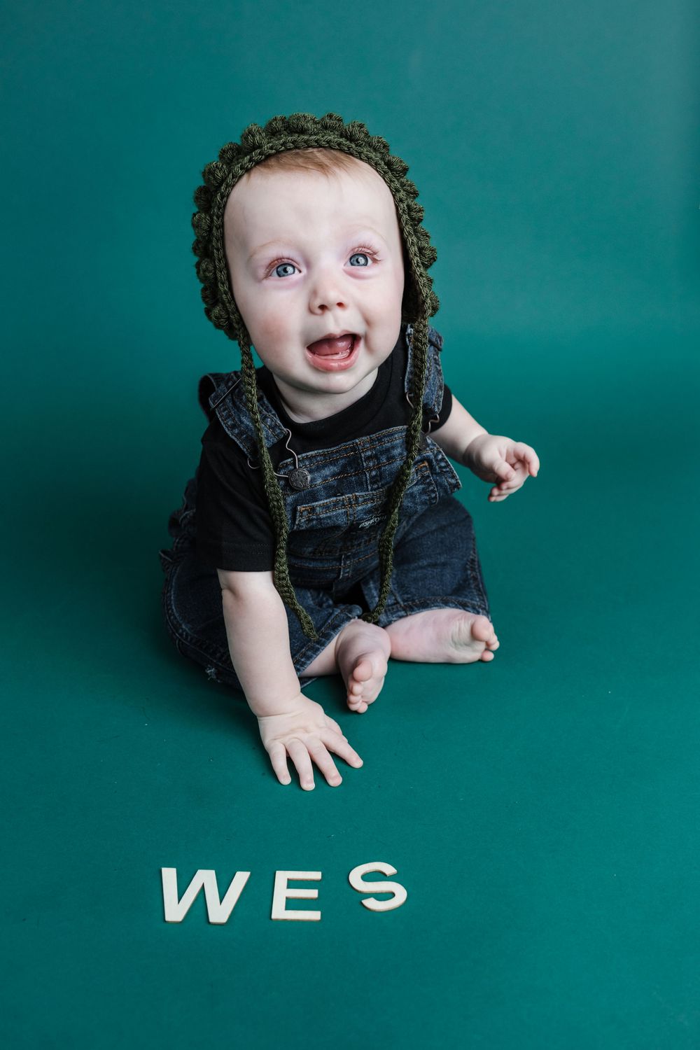 Small child wearing knit hat and denim overalls sits against teal backdrop with 'WES' text below.