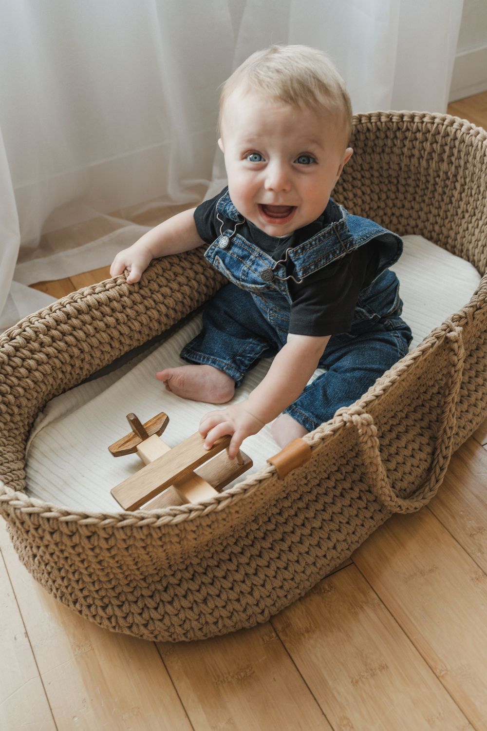 Baby in denim overalls sits in woven basket with wooden toys, smiling brightly against white curtain backdrop.