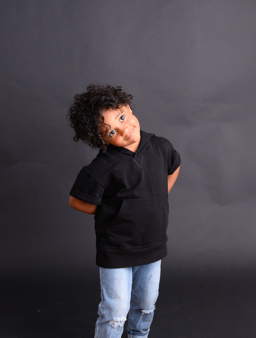 Playful portrait of child in black hoodie and ripped jeans against dark background.