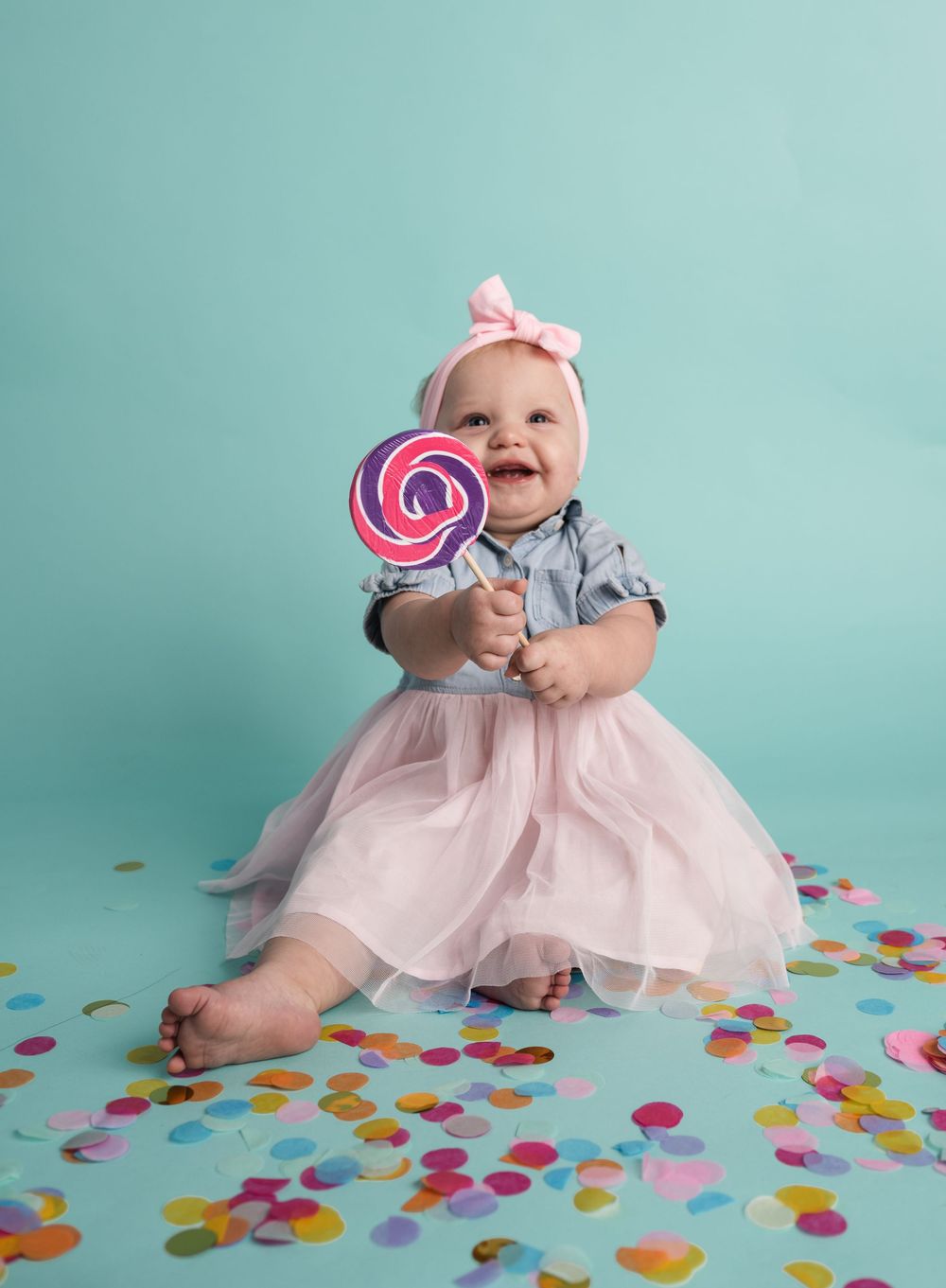 Baby in pink tulle dress plays with colorful lollipop and confetti on turquoise backdrop in milestone photo session.