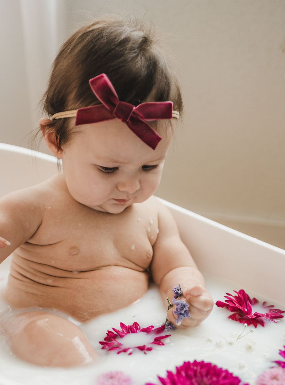 A baby with a red bow headband explores white bubbles and pink flowers during bath time.