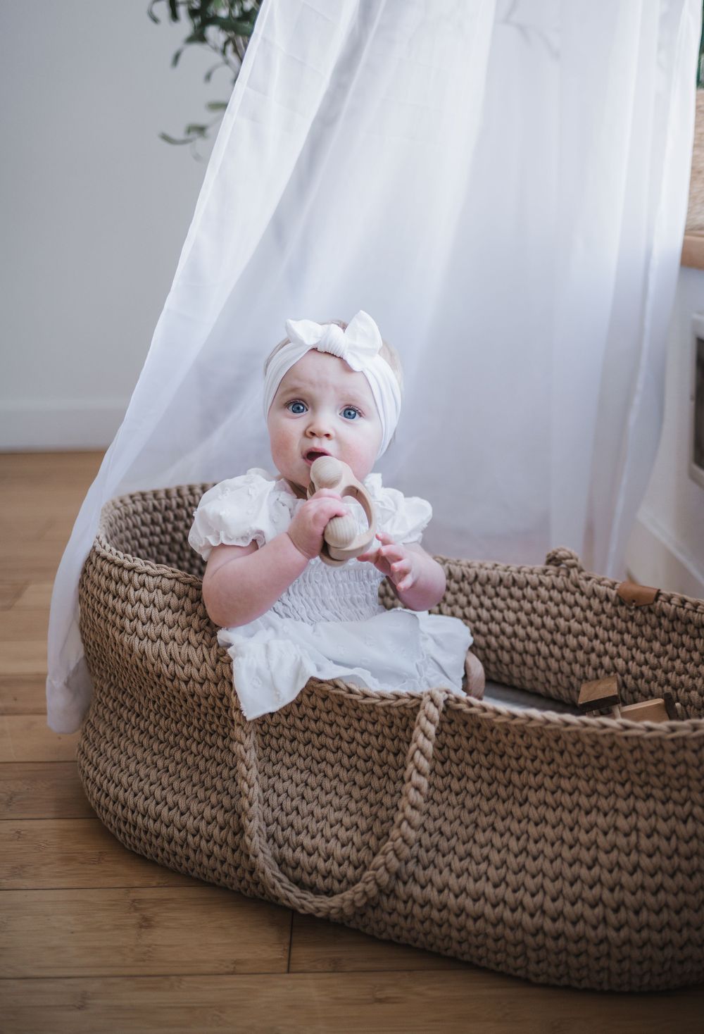 Baby in white dress sitting in large woven moses basket beside white curtain on hardwood floor.