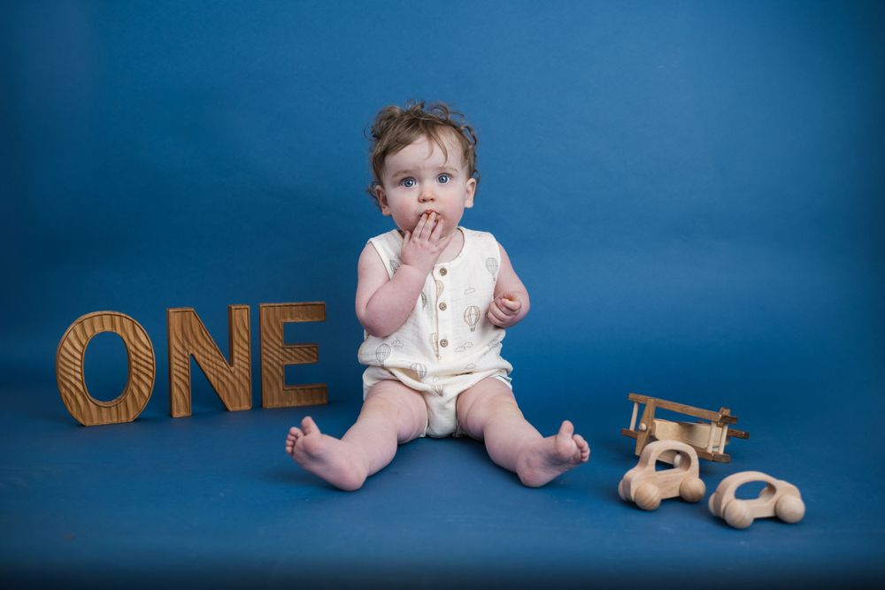 Baby sits against blue backdrop with wooden ONE text and teddy bear decorations.