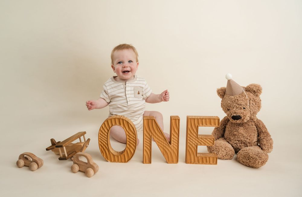 Baby's first birthday photo with wooden ONE letters, teddy bear with party hat, and toy airplane on cream background.