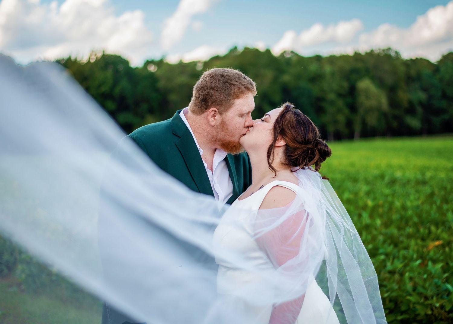 Romantic wedding portrait of couple kissing in field with flowing veil at sunset.