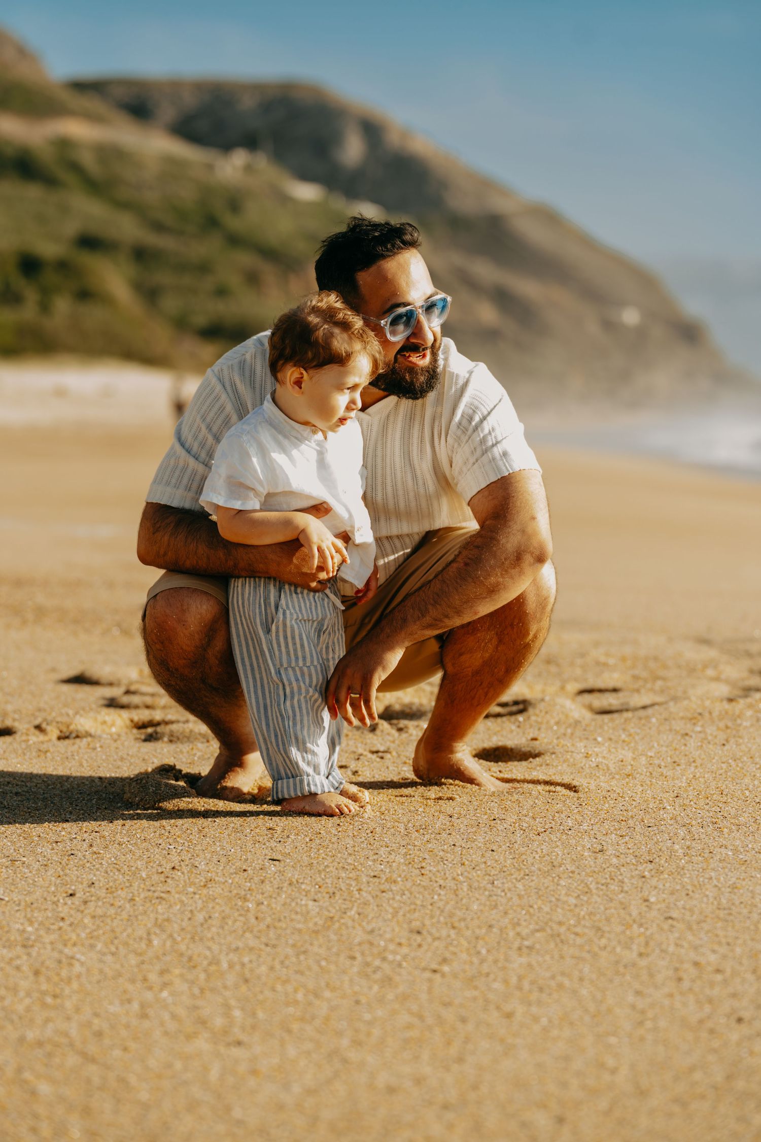 Two people sit close together on a sandy beach with mountains in the background during golden hour.