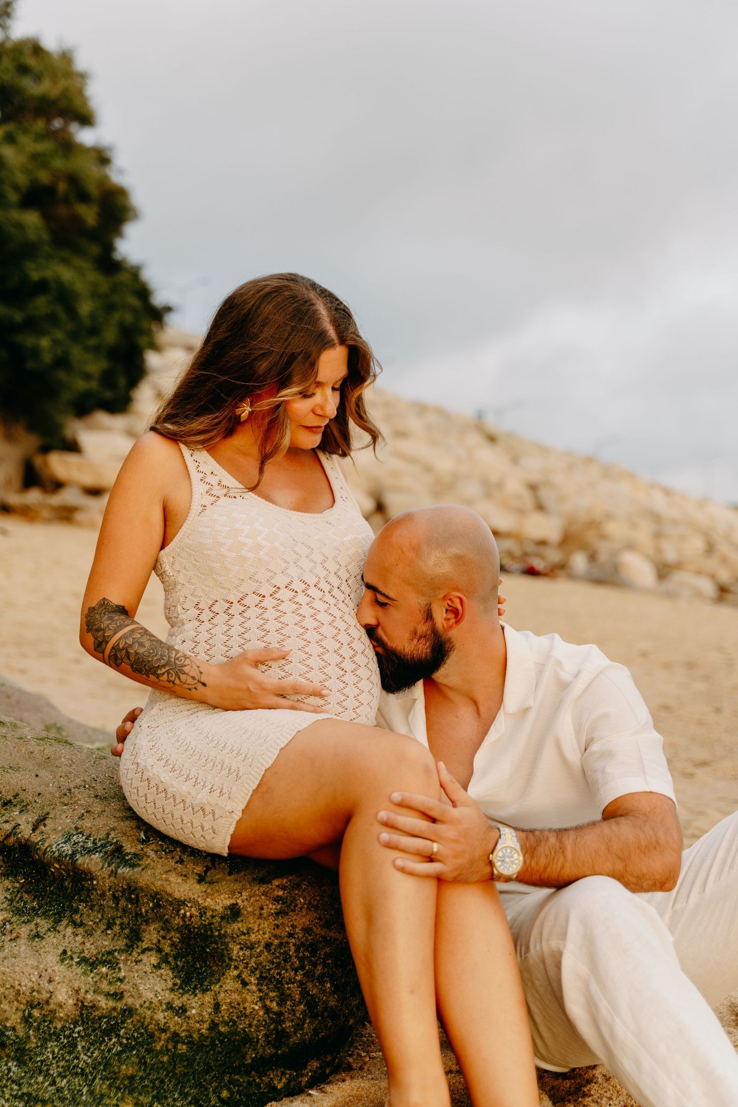 Romantic couple in white outfits sharing intimate moments on a rocky beach during sunset.