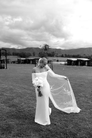 A graceful bride in a fitted off-shoulder gown holds up her delicate veil on a grassy field with mountains in the background.