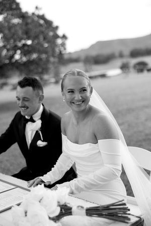 Black and white photo of a happy bride and groom sitting together at an outdoor wedding venue in a natural setting.