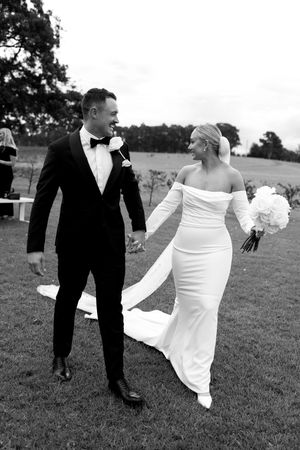 Black and white photo of newlyweds walking hand in hand on a grassy lawn, bride in sleek gown and groom in tuxedo.