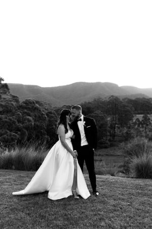 A romantic black and white wedding photo of a couple in formal attire embracing on a hillside with mountains in the background.